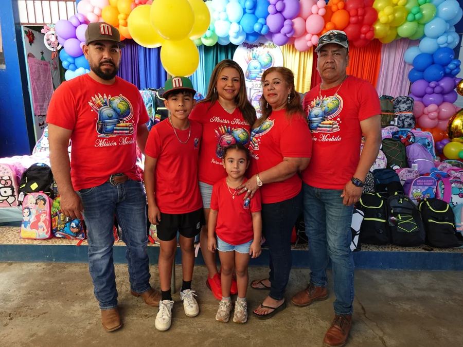 The Misión Mochila family team in matching red shirts beside stacks of backpacks