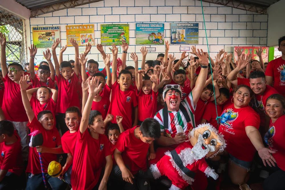 Children in red Misión Mochila shirts cheering with volunteers and a clown at a school celebration