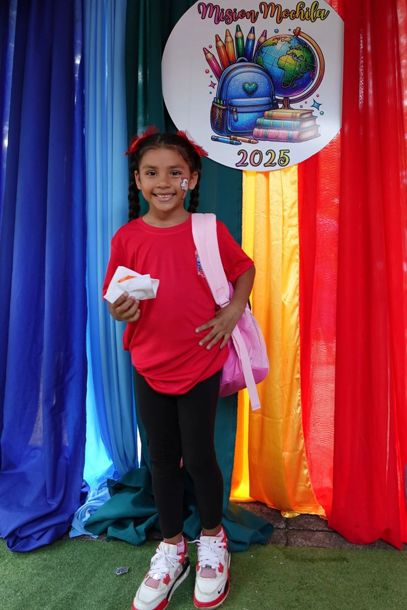 A smiling girl holds her new backpack in front of the rainbow Misión Mochila 2025 backdrop
